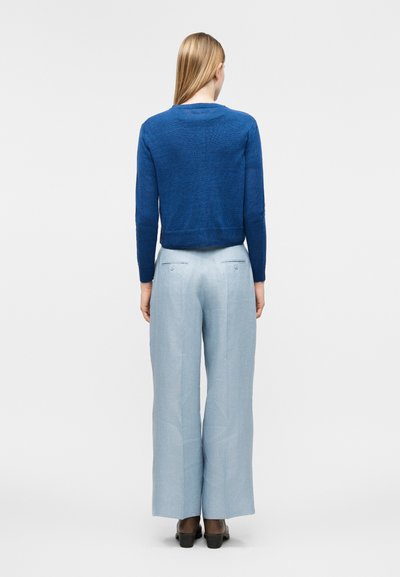 Άτομο που φοράει μπλε πουλόβερ με μακριά μανίκια, ανοιχτό μπλε παντελόνι φαρδιάς γραμμής και σκούρα καφέ παπούτσια, στέκεται γυρισμένο με την πλάτη σε απλό φόντο.