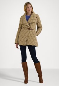 Casaco acolchoado bege com um colarinho largo, costura diagonal, cintura cingida por um cinto e detalhes em couro castanho, combinado com jeans escuros e botas castanhas.