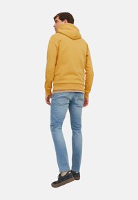 Gelbe Kapuzenjacke mit Kängurutasche, lässige Passform. Kombiniert mit hellblauen Slim-Jeans und schwarzen Schuhen mit braunen Sohlen. Schlichter Hintergrund.