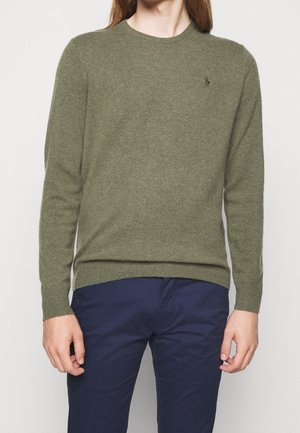 Man die een olijfgroene trui met lange mouwen en marineblauwe broek draagt, staand tegen een effen witte achtergrond.