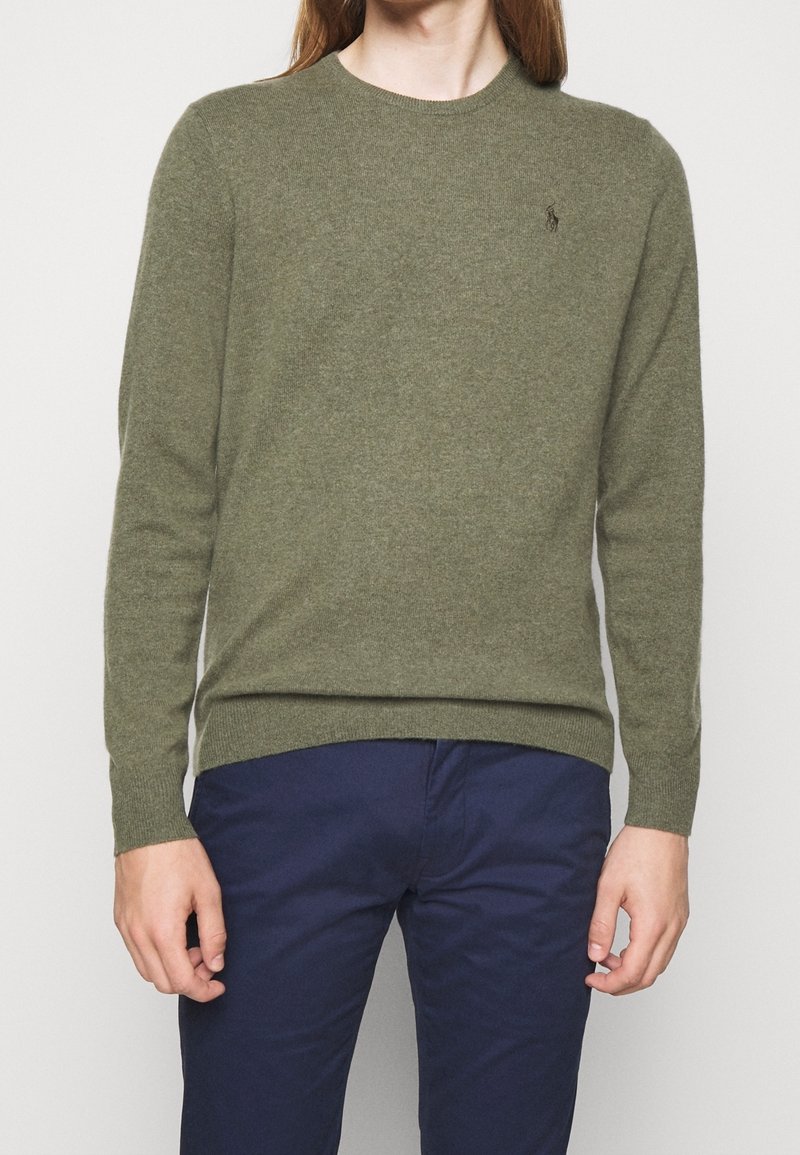 Homme portant un pull à manches longues vert olive et un pantalon bleu marine, debout devant un fond blanc uni.