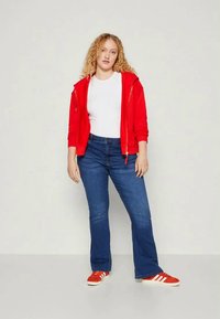 Fato de capuz vermelho com fecho, t-shirt branca, jeans azul de boca de sino e ténis vermelhos. Cabelo encaracolado; pose em pé contra um fundo neutro.