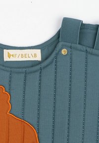 Kék, steppelt anyag narancssárga hímzéssel, amely egy sziluettet ábrázol. Állítható pántokkal és arany csattal rendelkezik. Felirat a tetején.