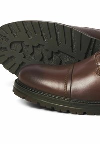 Bota de cuero marrón hasta el tobillo con punta redondeada, con suela de goma texturizada y detalles de costura visibles. La suela tiene un patrón de banda de rodadura resistente.