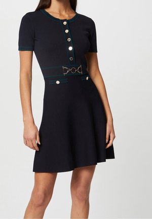 Marineblaues Kleid mit kurzen Ärmeln, silbernen Knöpfen vorne, grünem Besatz, dekorativem Gürtel mit Metallschnalle und zwei geknöpften Klappentaschen an der Taille.