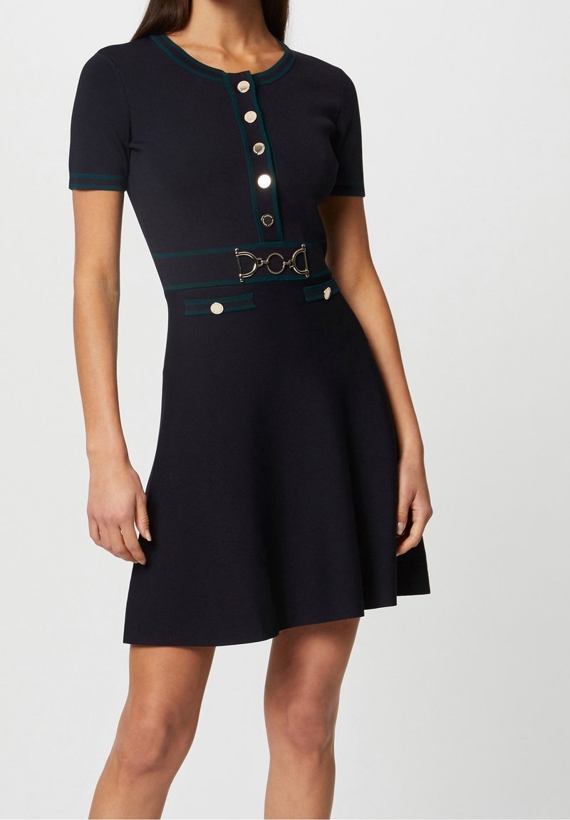 Robe bleu marine à manches courtes avec boutons argentés sur le devant, bordure verte, ceinture décorative avec boucle métallique et deux poches à rabat boutonnées à la taille.