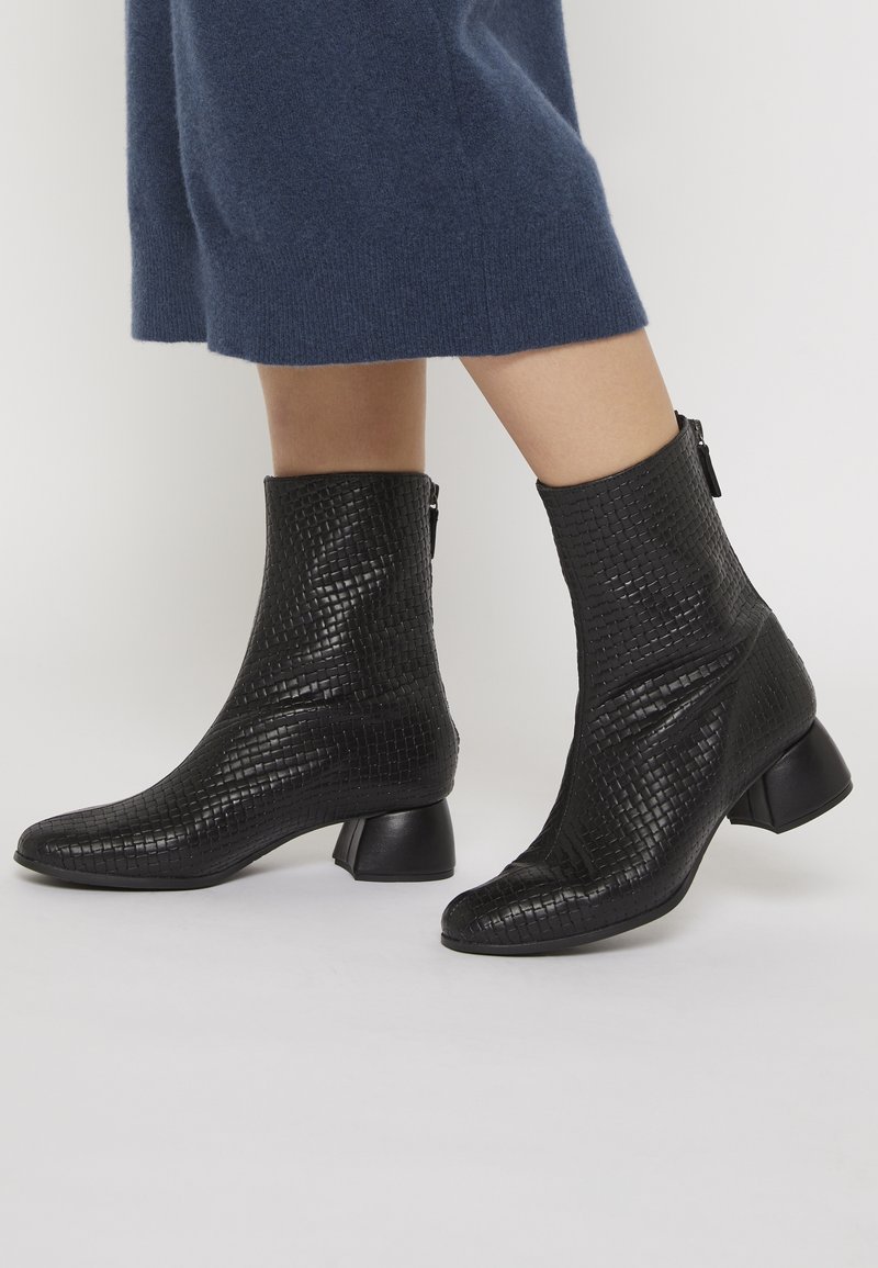 Bottes noires à la cheville avec un motif en relief texturé, bout pointu et petit talon carré. Détail de fermeture zipper sur le côté mis en avant.