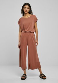 Rose-colored jumpsuit with short sleeves, elastic waistband, and wide-leg design. Features smooth texture and casual fit. Worn with black sandals.