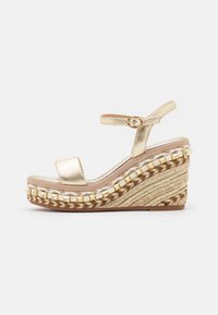 Sandalo wedge metallico dorato con cinturino alla caviglia, fascia anteriore ampia e suola in espadrille intrecciata con accenti bianchi e dorati.