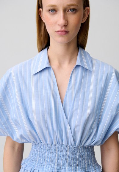 Giovane donna con capelli lisci castano chiaro che indossa una camicetta azzurra a righe bianche con scollo a V profondo e vita elasticizzata arricciata.