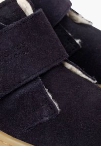 Tmavě námořnicky modré semišové boty s pásky na suchý zip, s měkkou bílou podšívkou a béžovou gumovou podrážkou. Detailní záběr zdůrazňuje texturu materiálu.