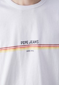 Fehér pamut póló, fekete "PEPE JEANS" felirattal, alatta piros, narancssárga és sárga vízszintes csíkokkal. Kerek nyakkivágás.