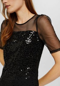 Černý flitrový top s průhlednými síťovinovými rukávy, kulatým výstřihem a texturovaným povrchem. Třpytivé flitry pokrývají přední stranu v gradientním vzoru.