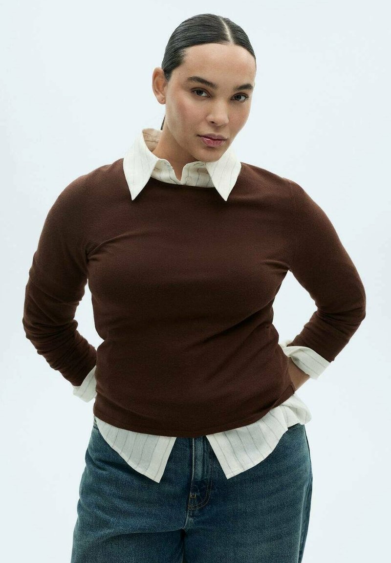 Maglione marrone a maniche lunghe indossato sopra una camicia bianca con colletto a strisce sottili. La modella indossa jeans di denim, posando con le mani sui fianchi.