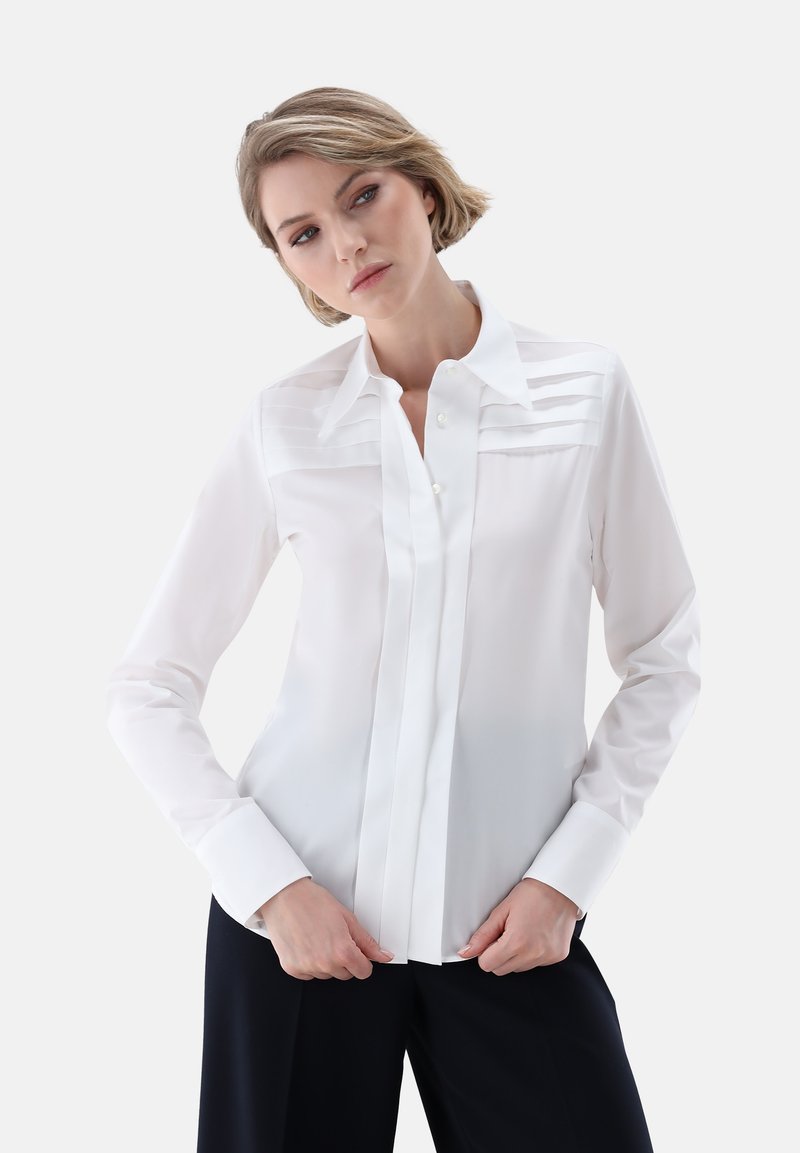 Camicia bianca a maniche lunghe con pieghe verticali sul petto, chiusura frontale con bottoni e collo classico. Tessuto morbido, vestibilità sartoriale.