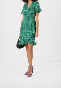 Robe portefeuille florale verte avec manches courtes, ourlet à volants et décolleté en V profond. Associée à des talons à brides violets et un sac à main noir.