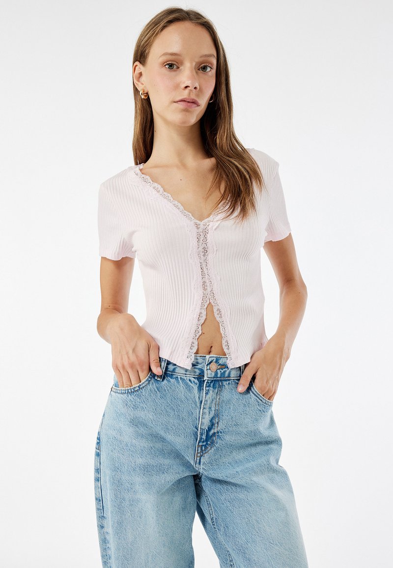 Helles rosa, geripptes Strickoberteil mit kurzen Ärmeln und Spitzenrand, V-Ausschnitt und Knopfverschluss, kombiniert mit hellblauen hochgeschnittenen Jeans.