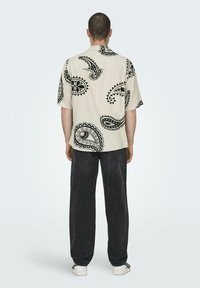 Beige Kurzarmhemd mit schwarzem Paisleymuster, lockere Passform und Rückenpasse. Kombiniert mit dunklen, weiten Hosen und weißen Sneakers.