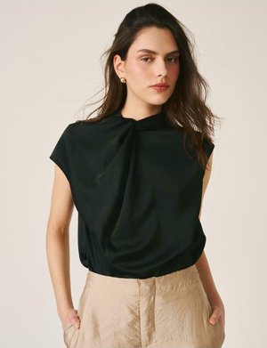 Mujer con cabello largo y oscuro, lleva pendientes dorados, una blusa negra sin mangas y pantalones beige de talle alto, con las manos en los bolsillos.