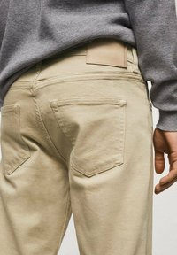 Pantalones de algodón beige con dos bolsillos traseros, detalles cosidos y una textura suave. Un suéter gris claro es parcialmente visible arriba.
