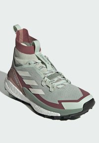 Vaaleanvihreä urheilukenkä, jossa on viininpunainen yksityiskohta, korkea neulottu varren osa, teksturoitu päällinen ja valkoinen välipohja, jossa on kolme valkoista raita sivussa.