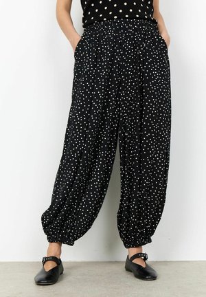 Pantaloni neri larghi con pois bianchi, caviglie elastiche, tasche, indossati con scarpe Mary Jane nere su un pavimento in cemento.