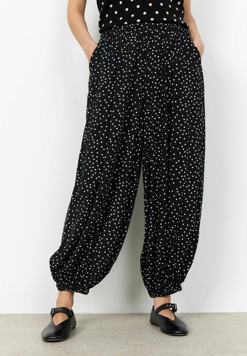 Pantalon noir ample à pois blancs, chevilles élastiques, poches, porté avec des chaussures Mary Jane noires sur un sol en béton.