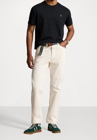 Černé bavlněné tričko, krémové kalhoty s texturovaným povrchem a zelené semišové tenisky s hnědými gumovými podrážkami. Pás s horizontálními pruhy.