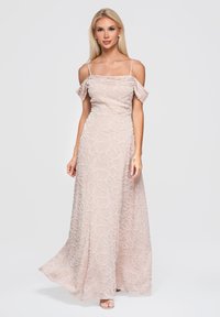 Femme portant une robe de soirée rose clair, épaules dénudées, avec une broderie florale, se tenant devant un fond blanc uni.