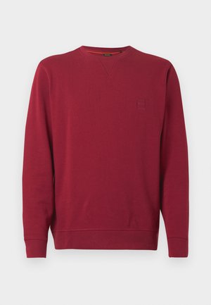 Czerwona bluza wykonana z miękkiego materiału, z okrągłym dekoltem, długimi rękawami oraz małą naszywką z logo na piersi. Gładka struktura na całej powierzchni.