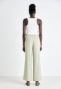 Femme aux cheveux tressés portant un débardeur blanc, un pantalon large vert clair et des sandales, debout, de dos, sur un fond uni.