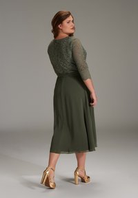 Olivegrünes Kleid mit dreiviertellangen Spitzenärmeln, A-Linienrock und glatter Oberfläche. Kombiniert mit goldenen High Heels.