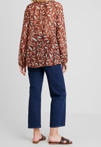 Blouse fluide semi-transparente marron avec un motif de feuilles, manches élastiques, associée à un denim foncé et des sandales brunes à lanières.