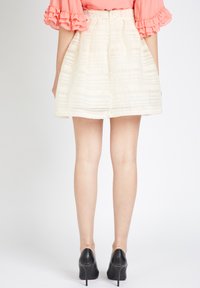 Mujer con una falda blanca texturizada, blusa de coral con volantes y tacones altos negros, de pie con la espalda hacia la cámara contra un fondo blanco.
