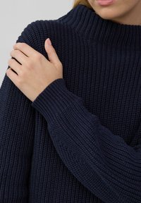 Navyblauwe gebreide trui met een hoge geribbelde kraag, textuurstof en geribbelde manchetten; close-up van schouder en arm.