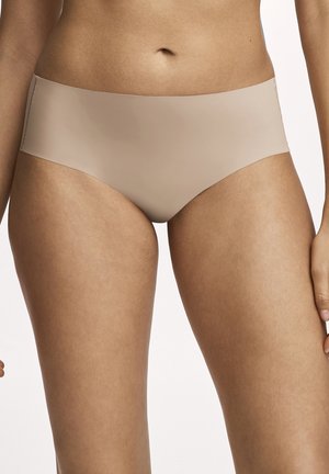 Frau trägt nahtlose beige Hipster-Unterwäsche, die den Mittelteil und die oberen Oberschenkel vor einem einfarbigen Hintergrund zeigt.