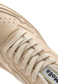 Beige sneaksi ar baltām austām šņorēm, perforētu pirkstu daļu un redzamu baltu birku ar melnu un dzeltenu tekstu uz mēles.