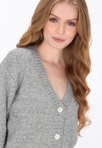 Szary dzianinowy kardigan z głębokim dekoltem w serek, trzema guzikami i teksturowanym wykończeniem z delikatnym blaskiem. Miękki, lekki materiał.