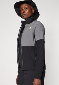 Melnā un pelēkā krāsā zipa jakā ar augstu apkakli, kas raksturojas ar strukturētu audumu, sānu kabat�ām un North Face logotipu uz krūšu daļas.