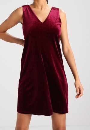 Bezrekawna sukienka z głębokiego czerwonego aksamitu z dekoltem w kształcie litery V, noszona przez osobę stojącą z jedną ręką na biodrze na jednolitym tle.