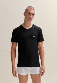 T-shirt en coton noir avec un col rond et des manches courtes, arborant un logo "BOSS" blanc sur la poitrine. Porté sur un short blanc.