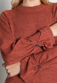 Rostfärgad stickad tröja med långa ärmar som har snodda manschetter och dekorativa knappdetaljer. Mjuk textur och rundad halsringning.