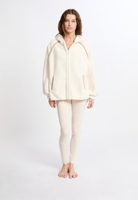 Giacca in pile con zip color crema e cappuccio, dotata di tasche laterali e cuciture marroni a contrasto; abbinata a leggings color crema, a piedi nudi.