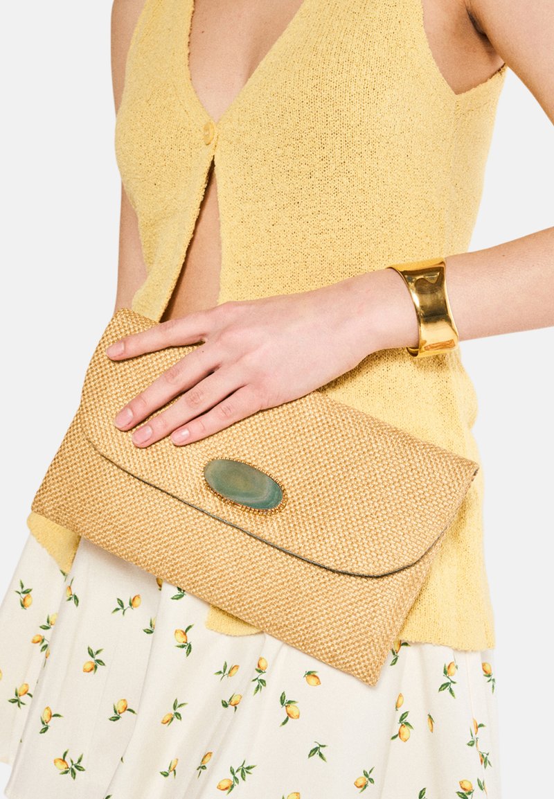 Vävd tan clutchväska med ett rundat grönt stenslå, hålls i en hand; parat med en gul stickad topp och mönstrad kjol.