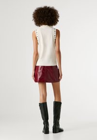 Göndör hajú személy, aki ujjatlan, csipkés fehér felsőt, bordó bőr mini szoknyát és fekete, térd fölé érő csizmát visel, háttal áll.