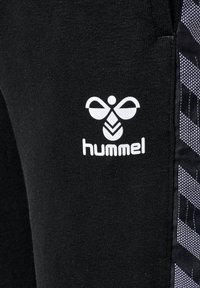 Schwarze Sporthosen mit einem weißen "hummel"-Logo. Mit einem strukturierten Streifenmuster an der Seite in Grau und Weiß. Dehnbarem Material.