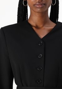 Robe noire avec un décolleté en V, ornée de quatre boutons noirs sur le devant, en tissu lisse et texturé. Cheveux tressés et boucles d'oreilles en créole visibles.