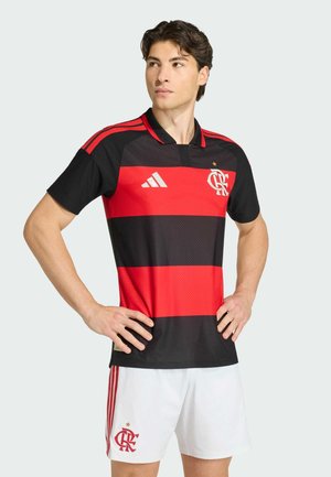 Jeune homme debout les mains sur les hanches, portant un maillot de football rayé noir et rouge et un short blanc avec un logo assorti sur fond clair.