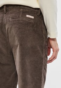 Pantaloni de corduroy maro, cu ribbing vertical, având un buzunar posterior și un mic etichete cu logo-ul Calvin Klein la talie.