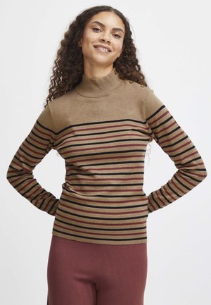 Donna sorridente con lunghi capelli ricci che indossa un maglione marrone a collo alto con strisce orizzontali nere e rosse e pantaloni color bordeaux, con le mani sui fianchi.